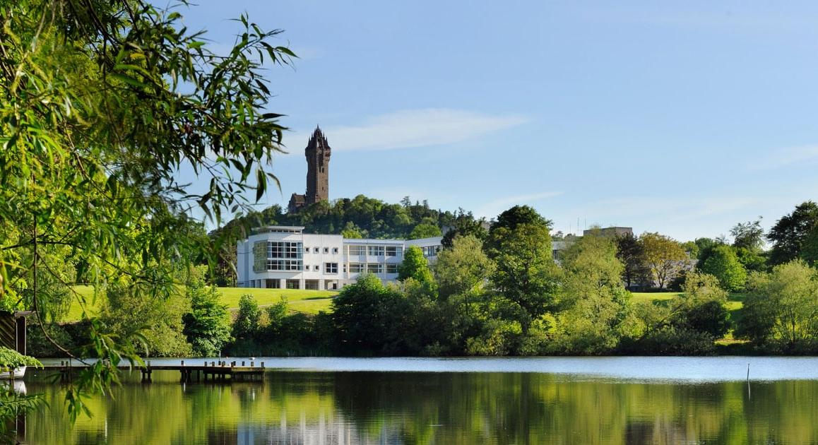 绿色的森林和平静的湖泊的照片, 后面有一座白色的建筑和一座高塔:斯特林的斯特林大学, Scotland.